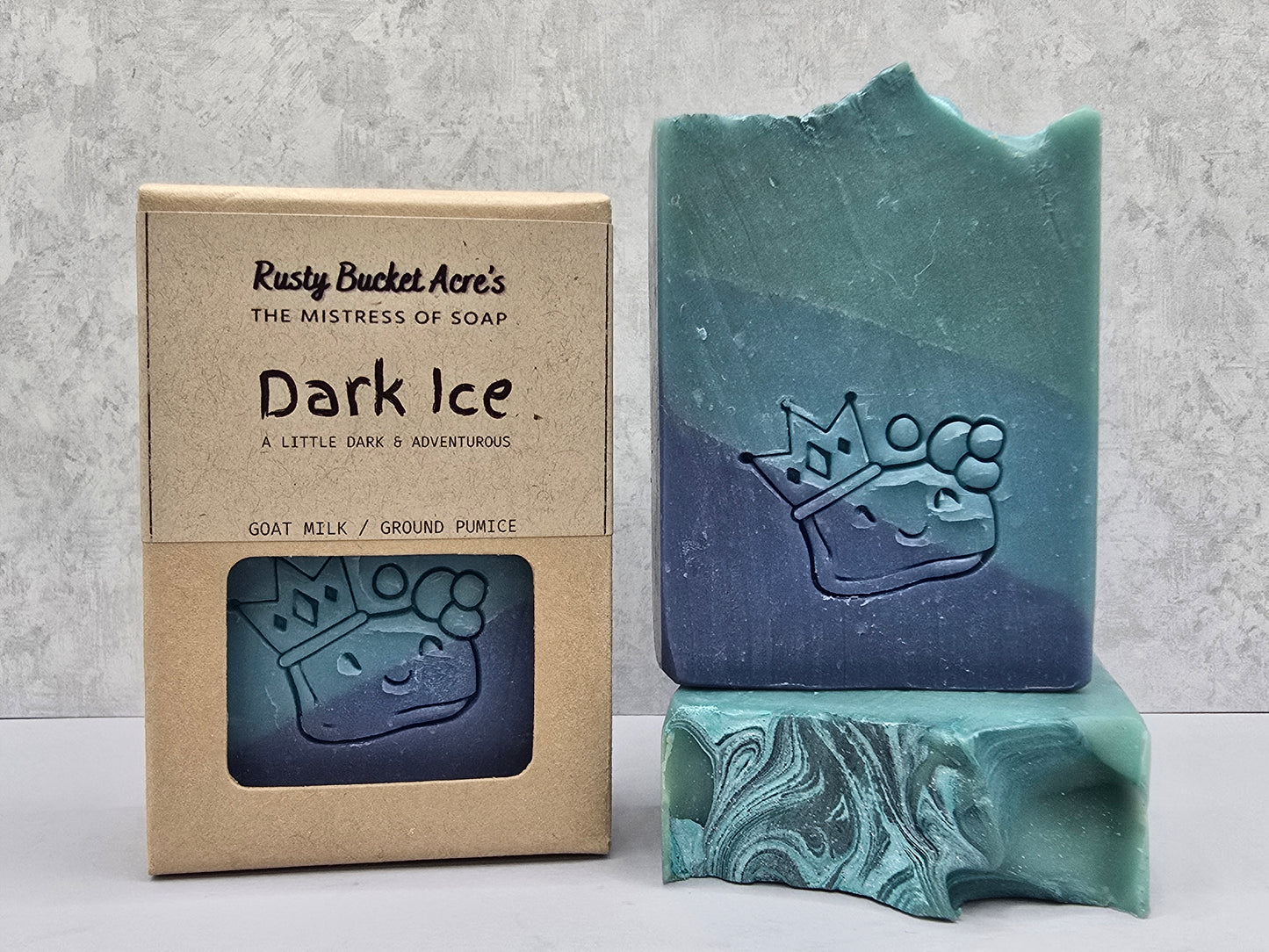 A bar of Rusty Bucket Acre's 'Dark Ice' goat milk soap shown next to its kraft paper packaging box and a decorative teal marble soap block, set against a textured gray background.