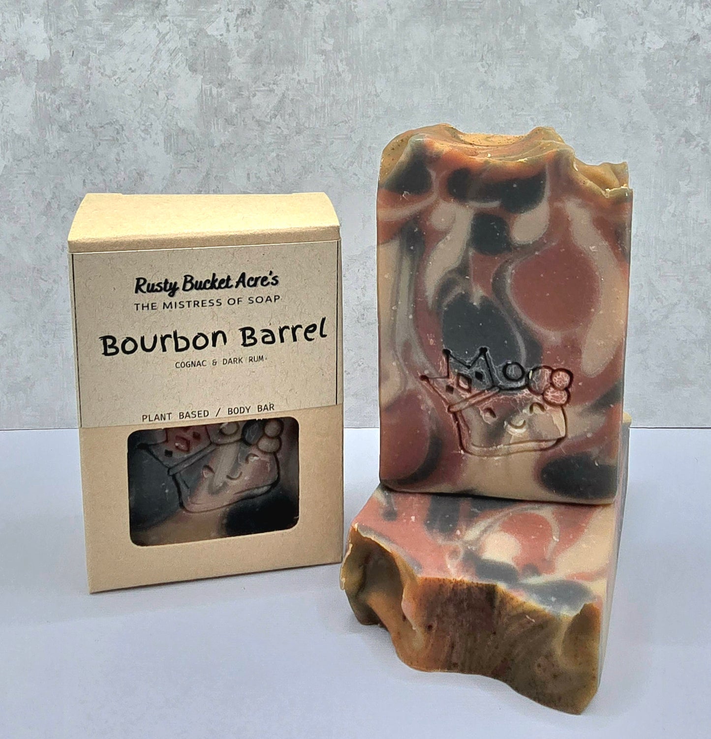 A handmade soap bar with a brown, beige, and black marbled camouflage pattern, shown next to its kraft paper packaging box labeled 'Rusty Bucket Acre's Bourbon Barrel'.