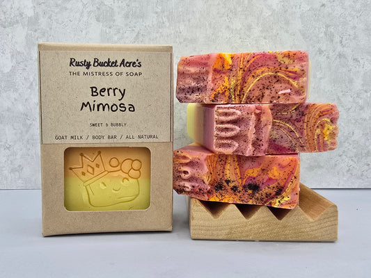 Three decorative handmade soap bars with pink, orange, and yellow swirls are stacked on a small wooden stand next to their kraft paper packaging box labeled 'Berry Mimosa' by Rusty Bucket Acre's.