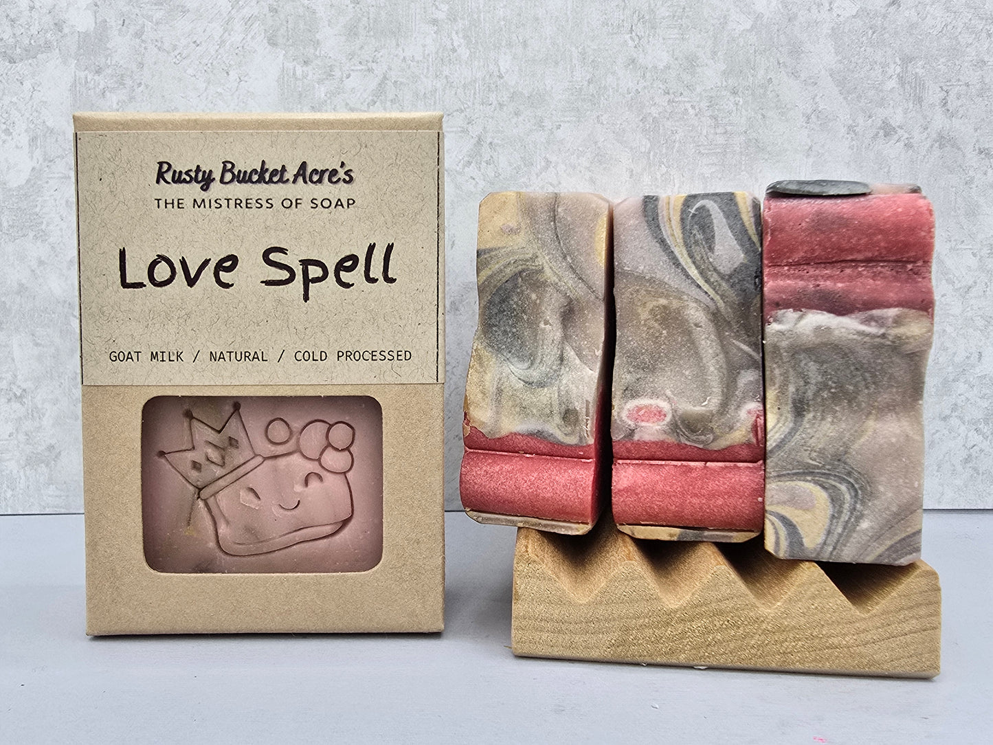 Three handmade soap bars with a pink, gray, and white marbled pattern are displayed next to their kraft paper packaging box labeled 'Love Spell' by Rusty Bucket Acre's.