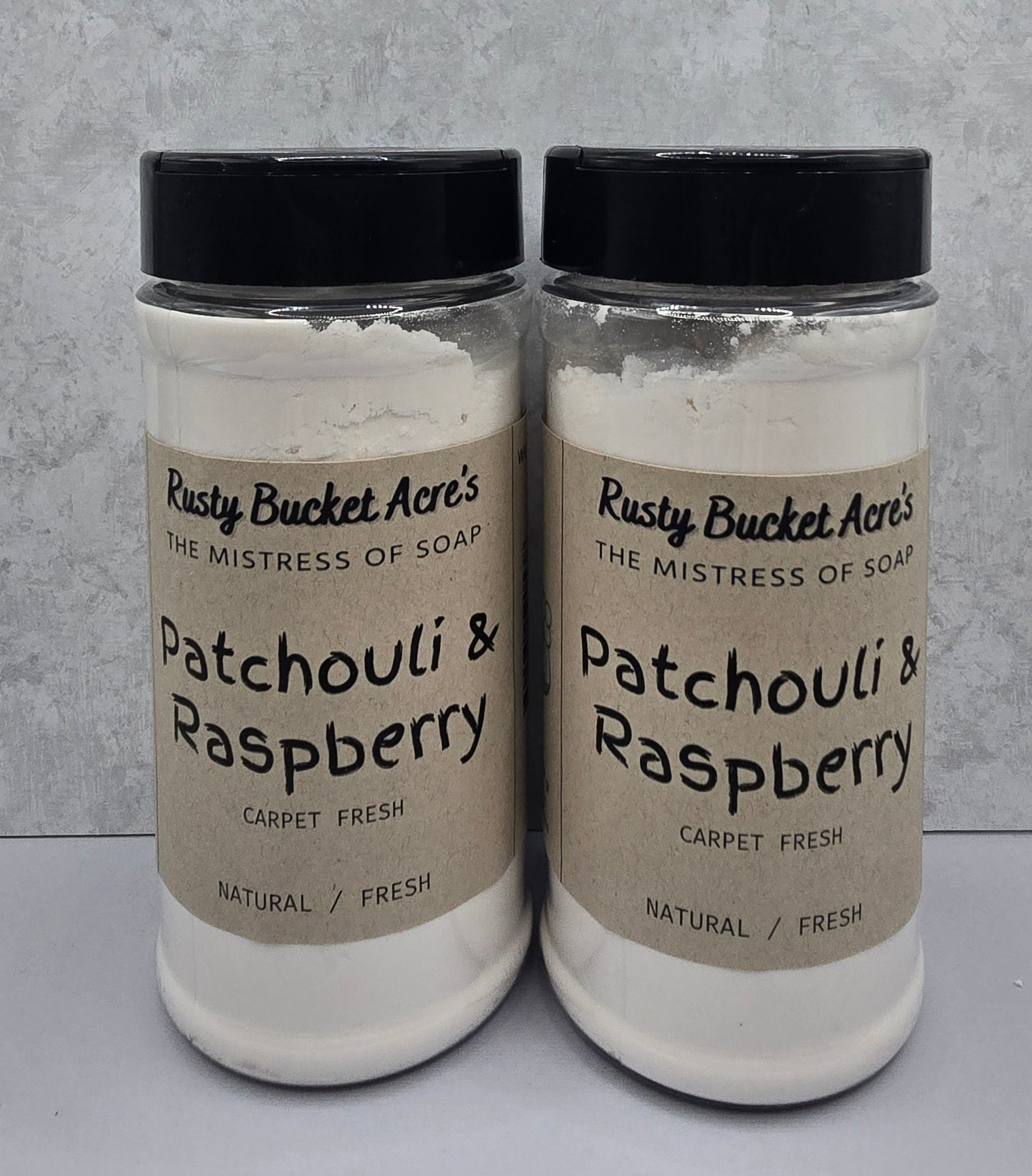 Two clear plastic jars of Rusty Bucket Acre's Patchouli & Raspberry Carpet Fresh soap powder, featuring beige labels and black lids, set against a light gray textured background.