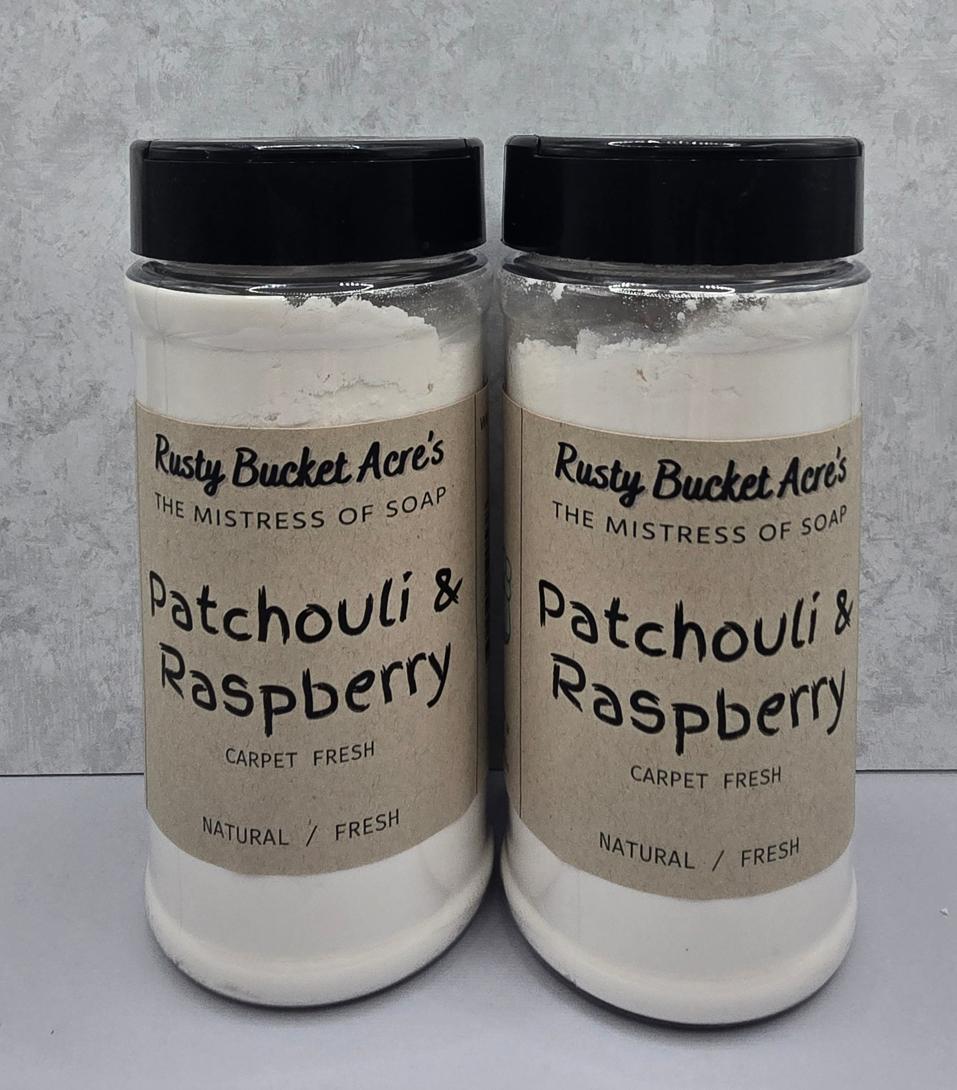 Two clear plastic jars of Rusty Bucket Acre's Patchouli & Raspberry Carpet Fresh soap powder, featuring beige labels and black lids, set against a light gray textured background.