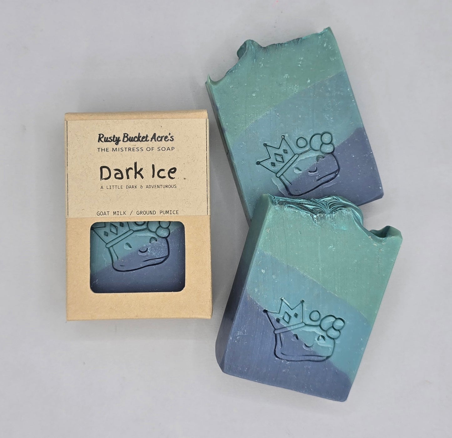 Two handmade bar soaps with a blue-green gradient and an embossed crown design, shown next to their kraft paper packaging box labeled 'Rusty Bucket Acre's Dark Ice'.