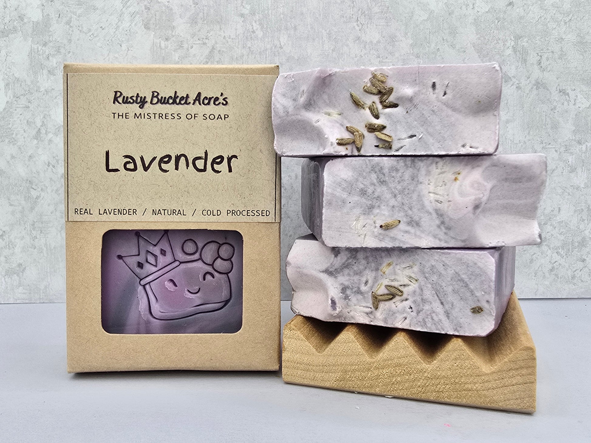 Lavender handcrafted soap bars with visible lavender buds, shown next to their kraft paper packaging box labeled 'Rusty Bucket Acre's Lavender'.
