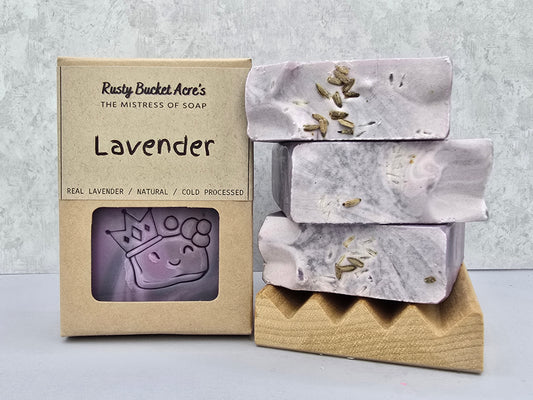 Lavender handcrafted soap bars with visible lavender buds, shown next to their kraft paper packaging box labeled 'Rusty Bucket Acre's Lavender'.