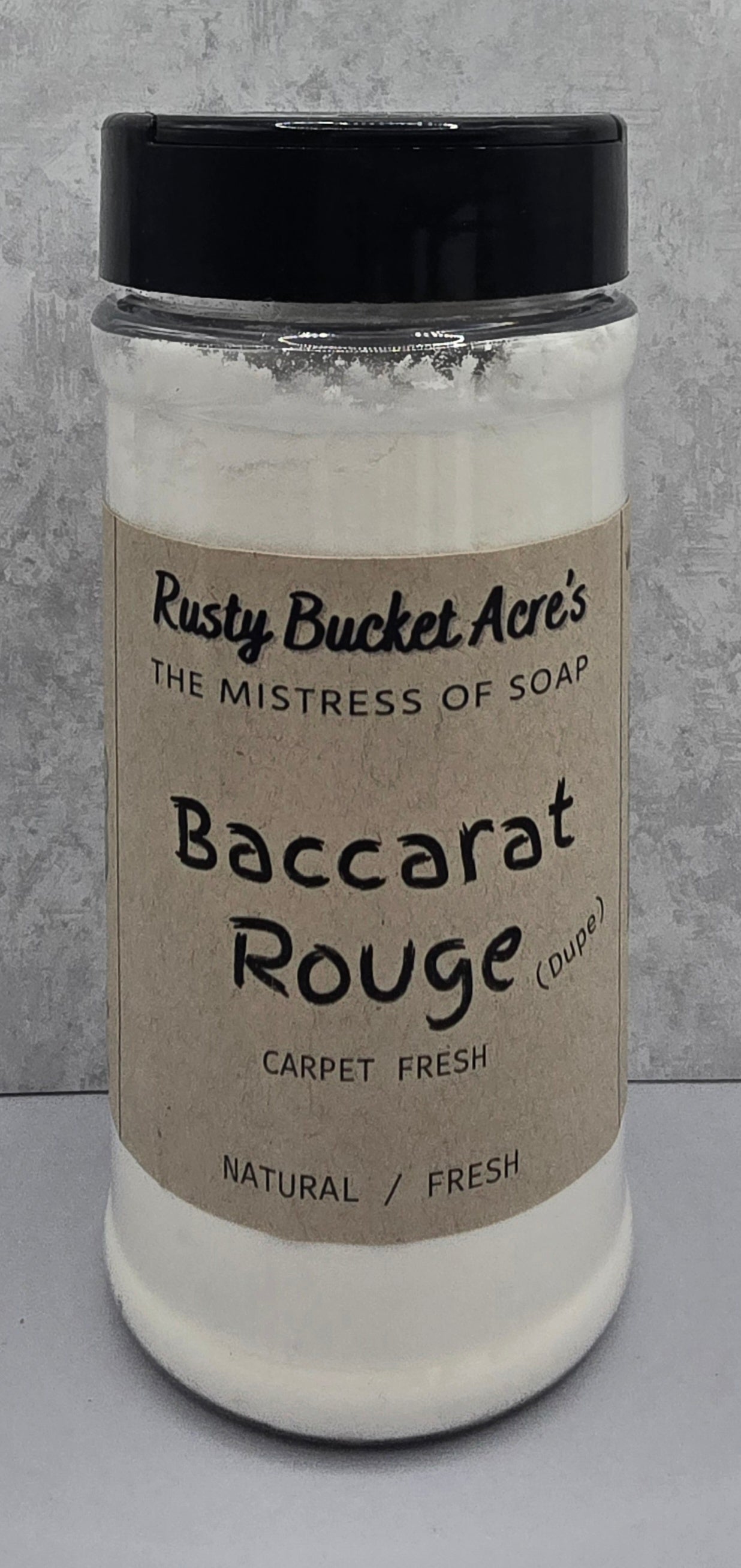 A clear plastic jar of Rusty Bucket Acre's 'Baccarat Rouge Dupe' carpet fresh soap, filled with white lather and topped with a black lid, set against a gray textured background. 