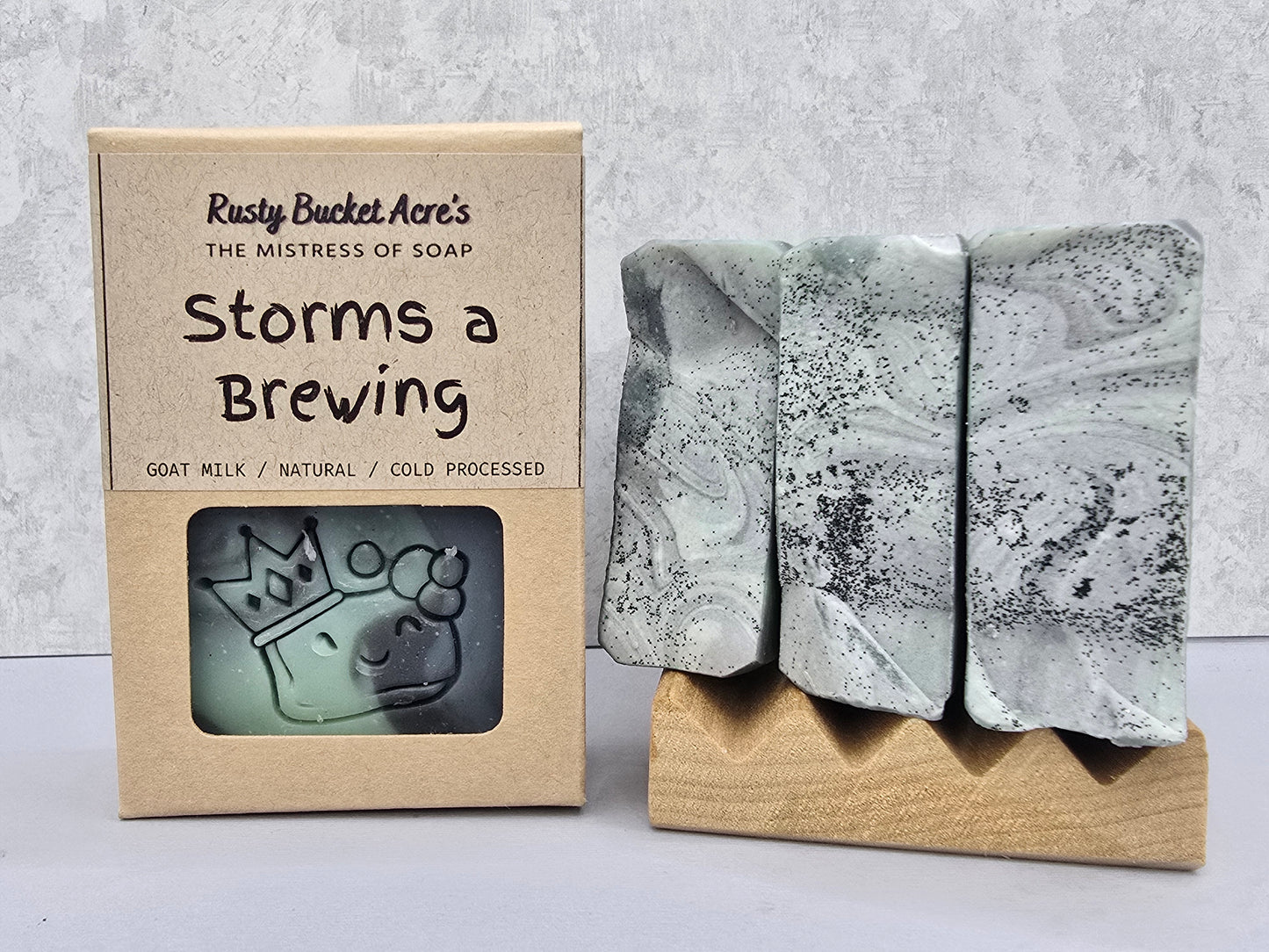 Three rectangular artisan soap bars with a gray and black marbled pattern displayed on a small wooden stand next to their kraft paper packaging box labeled 'Storms a Brewing' by Rusty Bucket Acre's.