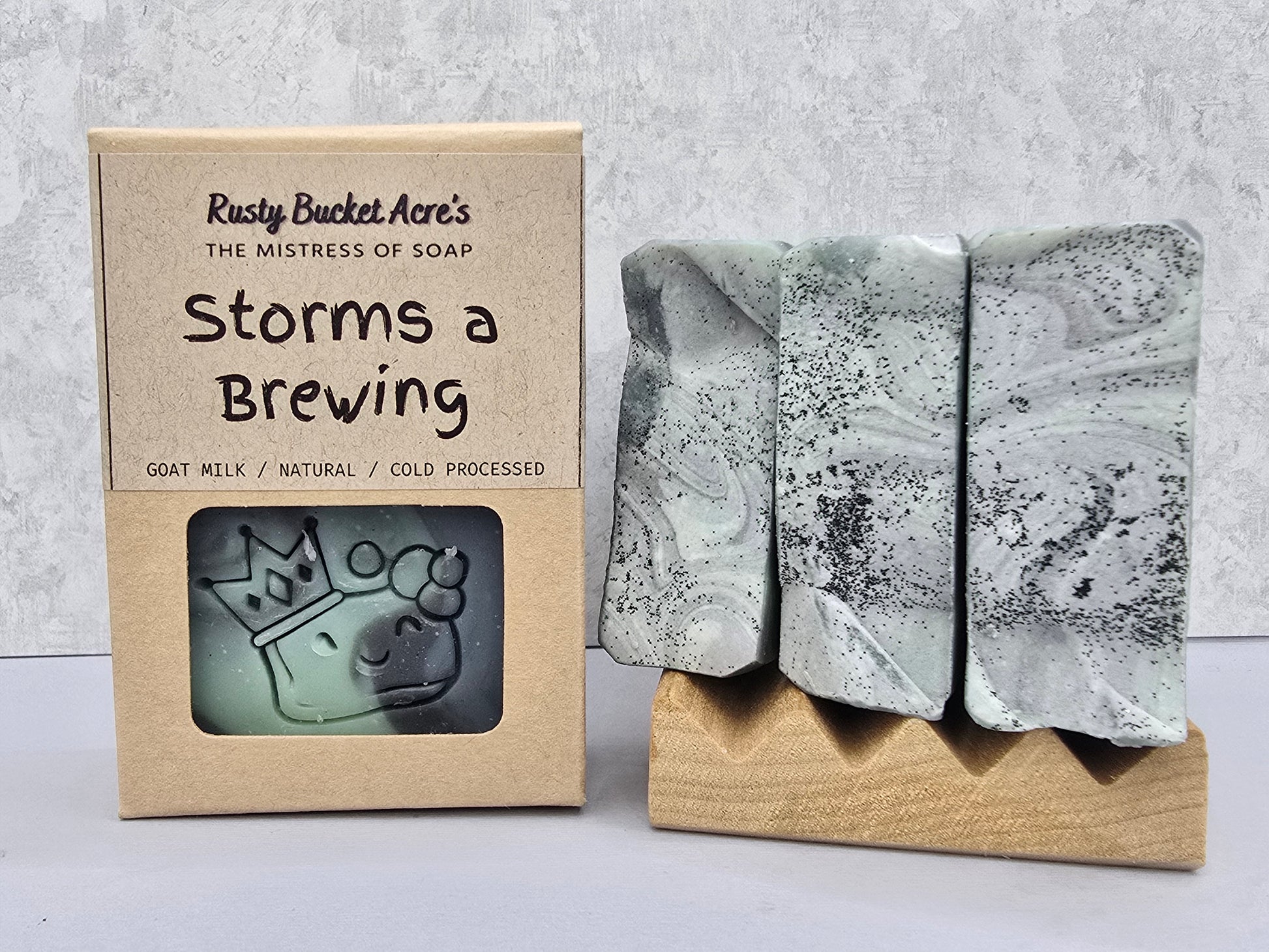 Three rectangular artisan soap bars with a gray and black marbled pattern displayed on a small wooden stand next to their kraft paper packaging box labeled 'Storms a Brewing' by Rusty Bucket Acre's.
