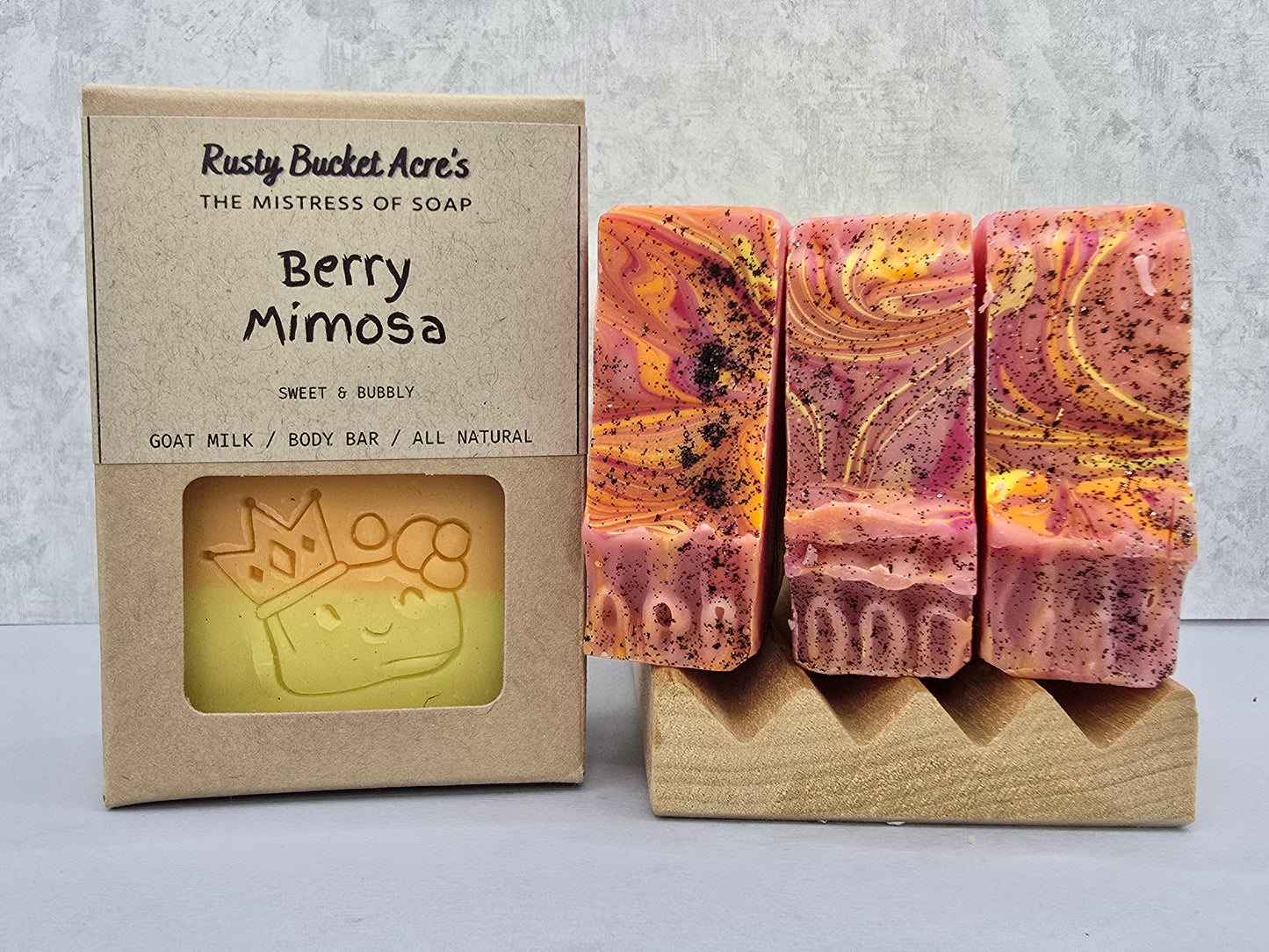 Three decorative soap bars with pink, purple, orange, and gold marbling are displayed on a small wooden stand next to their kraft paper packaging box labeled 'Berry Mimosa' by Rusty Bucket Acre's.