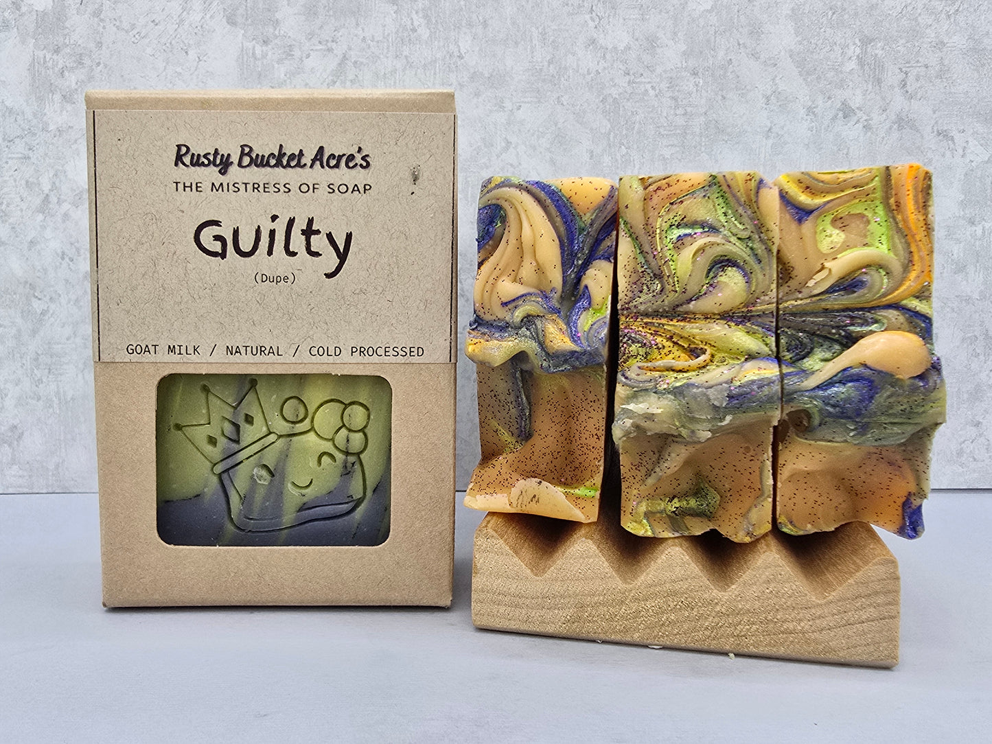 A handmade soap bar with a blue, green, and tan marbled swirl pattern displayed on a wooden stand next to its kraft paper packaging box labeled 'Rusty Bucket Acre's Guilty (Sour)' on a light gray background.