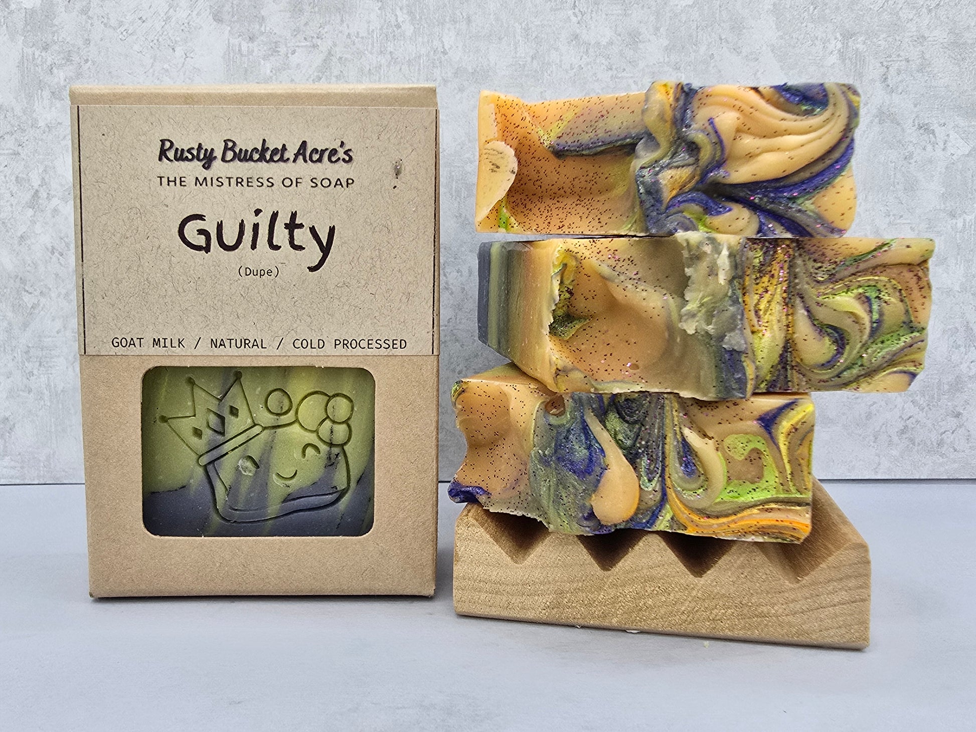 Three marbled soap bars with yellow, blue, and green swirls displayed on a wooden stand next to their kraft paper packaging box by Rusty Bucket Acre's.
