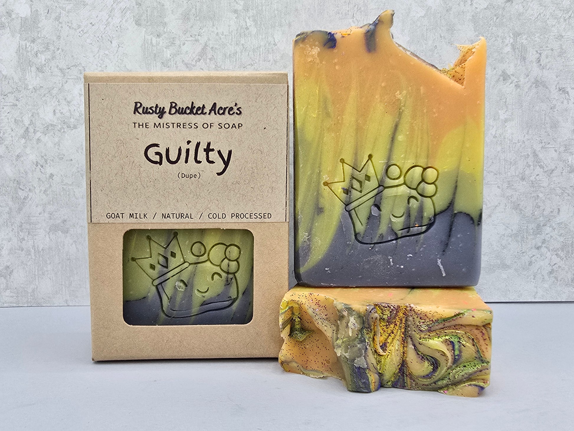 A handmade soap bar with a marbled yellow, green, and dark gray color pattern, shown next to its kraft paper packaging box labeled 'Rusty Bucket Acre's The Mistress of Soap Guilty'.