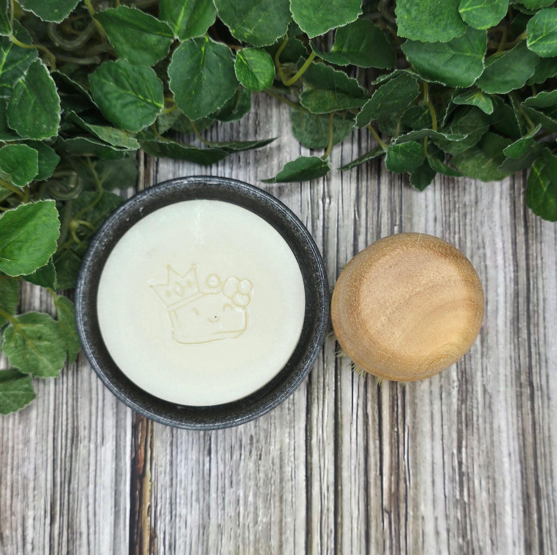 Bar of soap in a black container with a wooden lid on a wooden surface with green leaves.