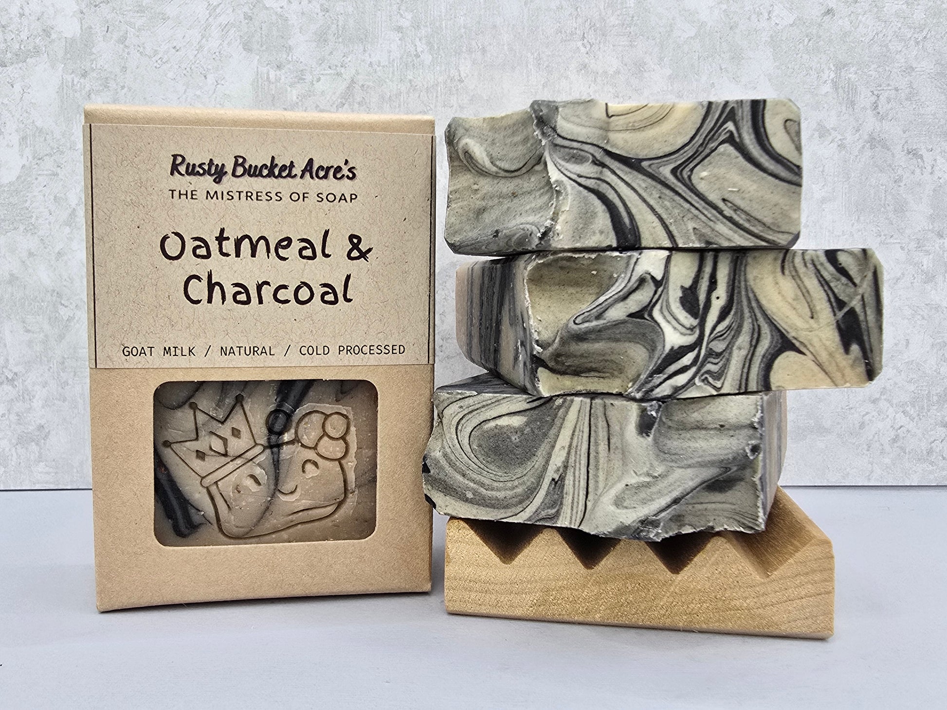 Three stacked rectangular bars of soap with a black, gray, and off-white marbled pattern displayed next to their kraft paper packaging box labeled 'Rusty Bucket Acre's Oatmeal & Charcoal'.