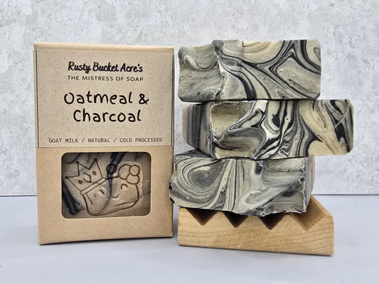 Three stacked rectangular bars of soap with a black, gray, and off-white marbled pattern displayed next to their kraft paper packaging box labeled 'Rusty Bucket Acre's Oatmeal & Charcoal'.