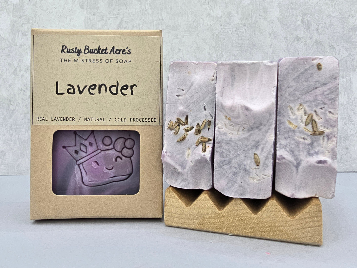 A handmade lavender soap bar with visible botanical inclusions, displayed next to its kraft paper packaging box labeled 'Rusty Bucket Acre's Lavender'.