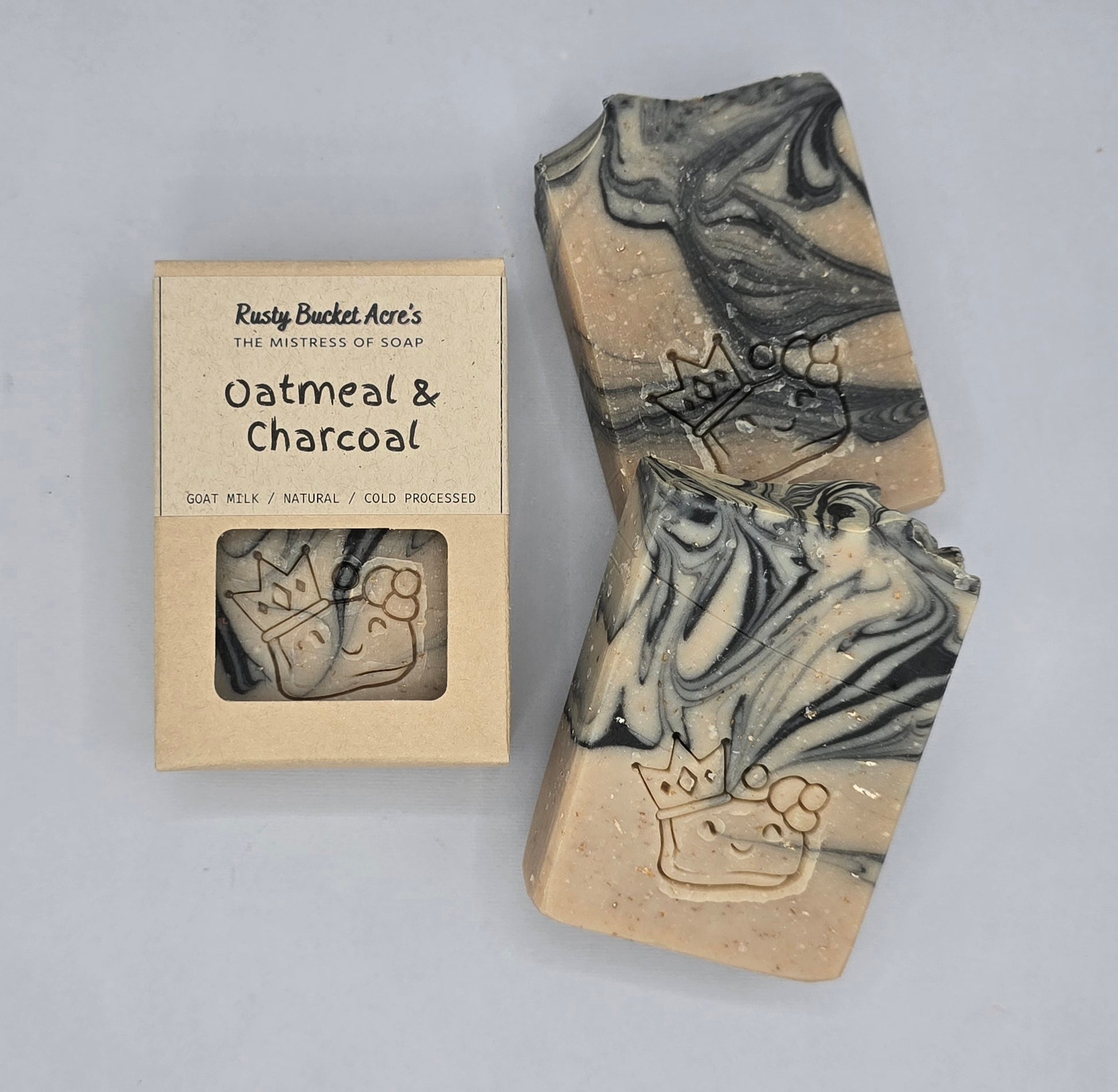 Two rectangular artisan soap bars with a marbled oatmeal and charcoal pattern and a carved crown design, displayed next to their kraft paper packaging box labeled 'Rusty Bucket Acre's Oatmeal & Charcoal'.