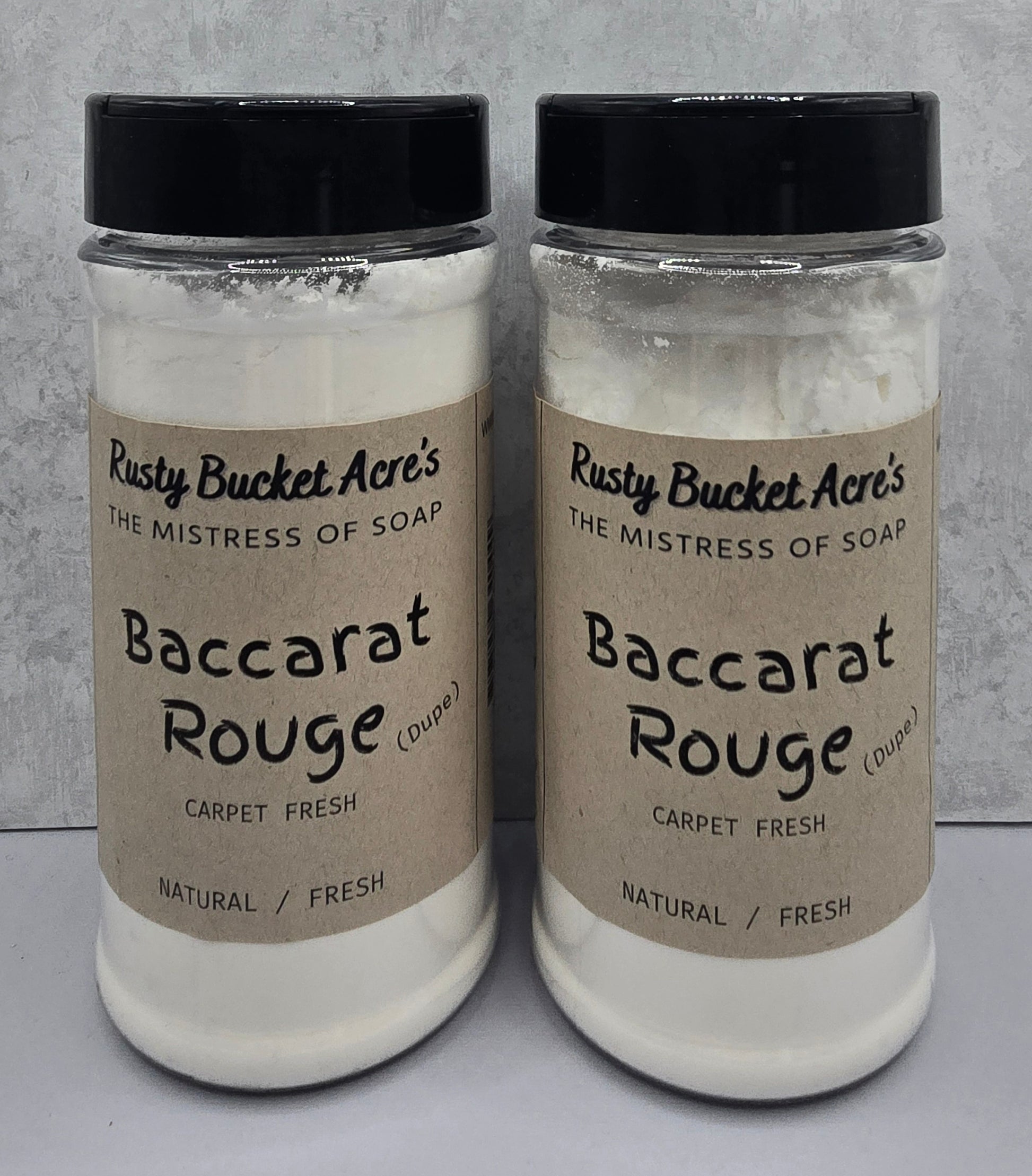 Two clear plastic jars of Rusty Bucket Acre's Baccarat Rouge (Dupe) Carpet Fresh natural fresh soap, filled with white soap powder and capped with black lids, set against a gray textured background.