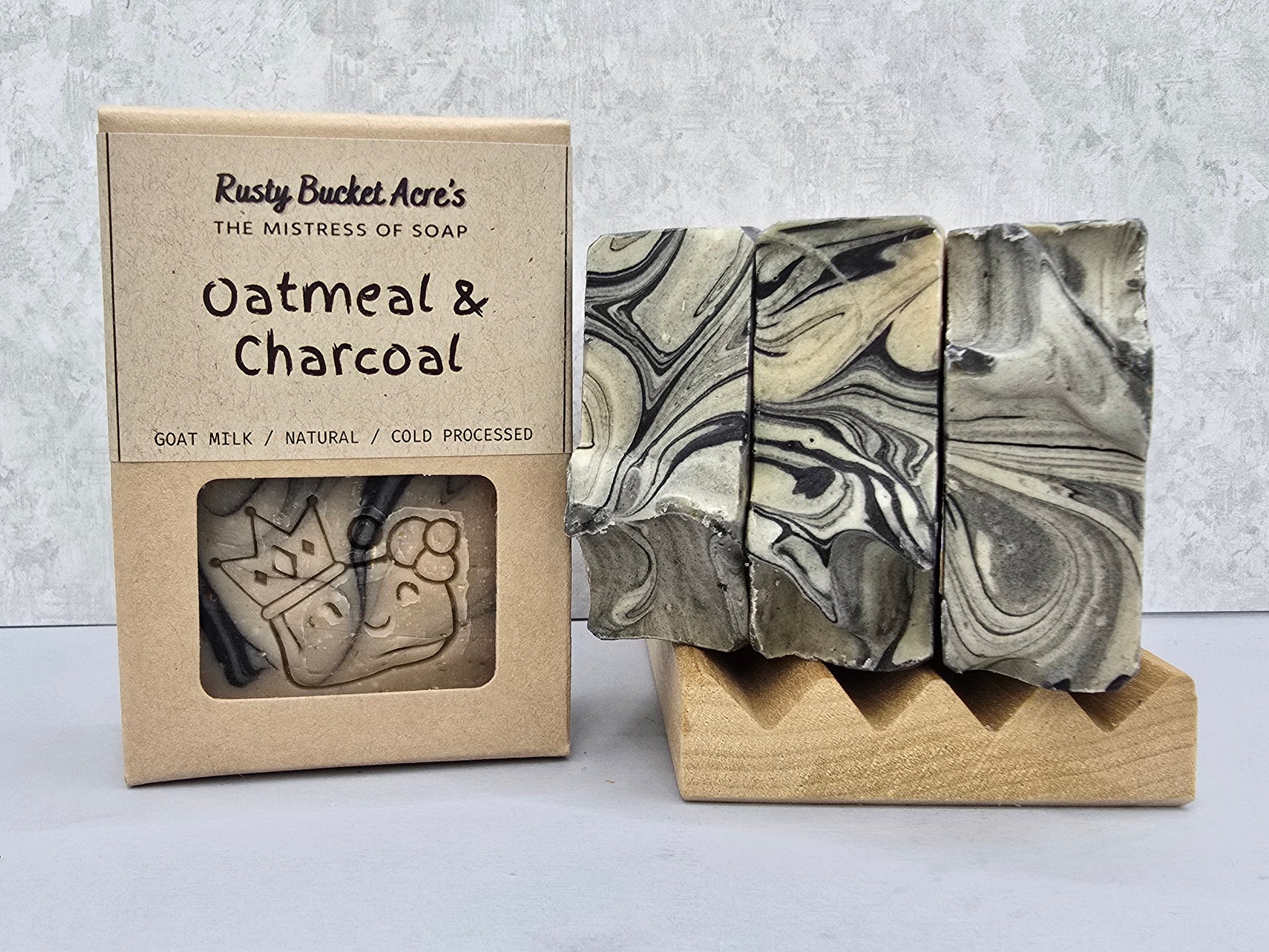 A bar of Rusty Bucket Acre's Oatmeal & Charcoal soap displayed next to its kraft paper packaging box on a light gray background.
