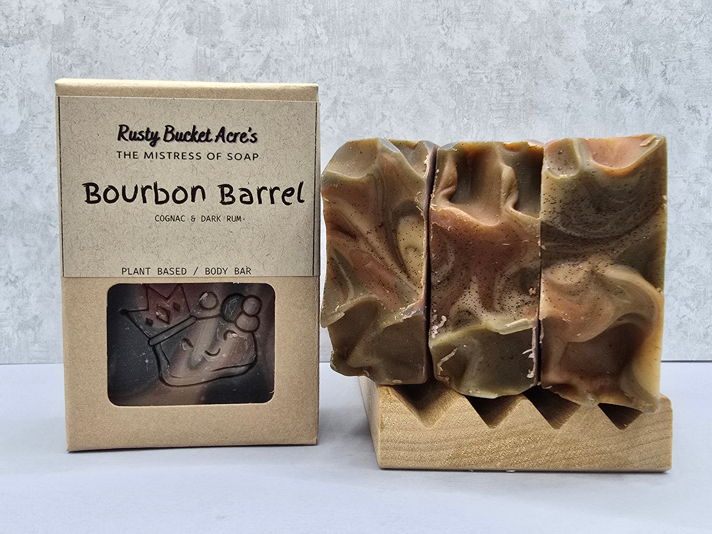A boxed body bar soap next to three carved bars of the same soap displayed on a wooden stand. The box reads 'Rusty Bucket Acre's The Mistress of Soap Bourbon Barrel' and shows a window with a soap bar inside.