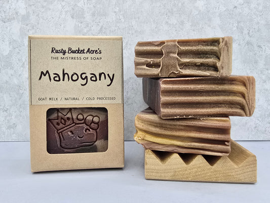 Mahogany scented soap bars displayed next to their kraft paper packaging box by Rusty Bucket Acre's. The soaps are stacked on a wooden stand, showing a marbled brown and beige texture, with the embossed brand logo 'RBAC' visible on the top bar.