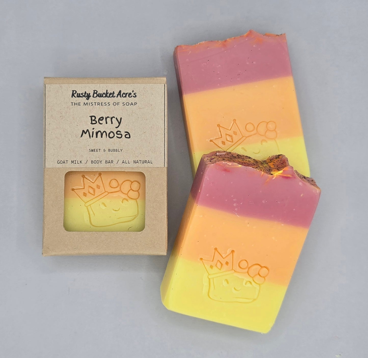 A handmade soap bar with pink, orange, and yellow horizontal layers next to its kraft paper packaging box. The box is labeled 'Berry Mimosa' by 'Raspy Bucket Acre's'.
