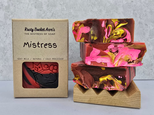 A boxed bar of soap and the stacked soap bars themselves on a small wooden stand. The packaging reads 'Rusty Bucket Acre's The Mistress of Soap Mistress' and shows a window with a red-and-black soap bar visible.