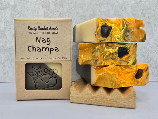 Three stacked handmade soap bars with gold and cream marbling and black stone inclusions, displayed next to their kraft paper packaging box labeled 'Rusty Bucket Acre's The Mistress of Soap Nag Champa'.