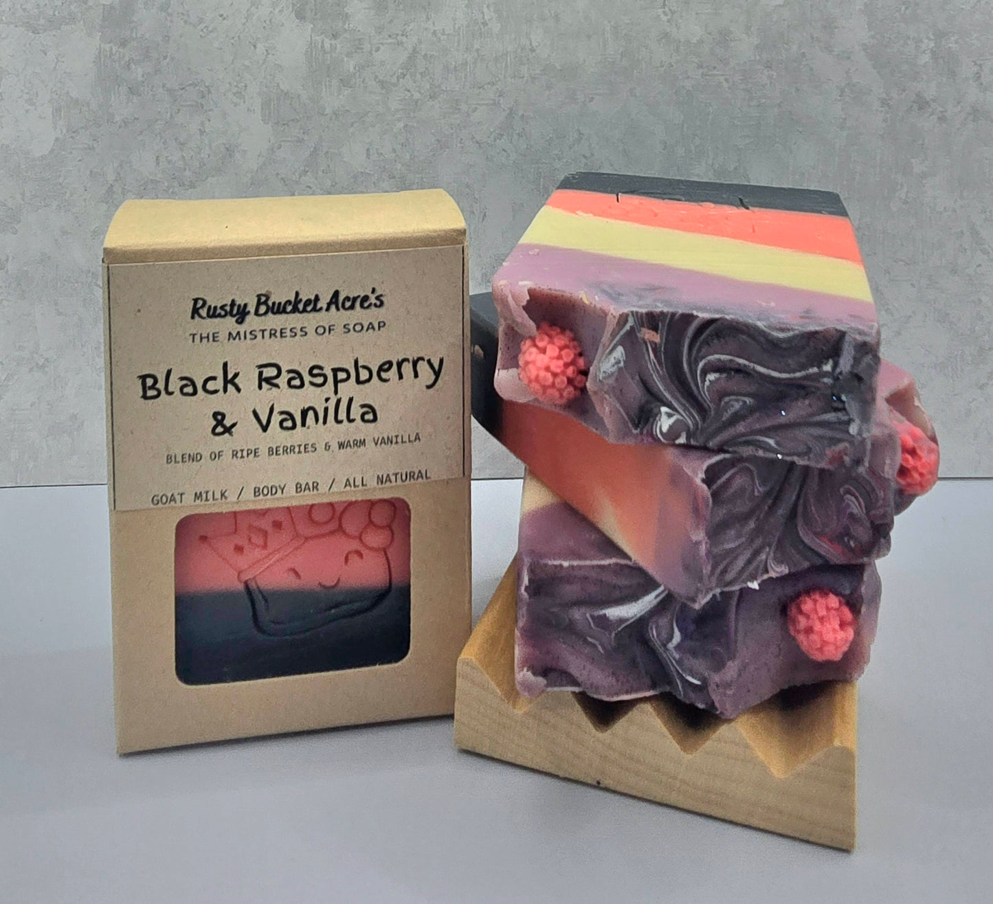 A handmade soap bar with layered black, purple, pink, and orange stripes, decorated with three pink raspberry charms, displayed next to its kraft paper packaging box labeled 'Rusty Bucket Acre's Black Raspberry & Vanilla'.