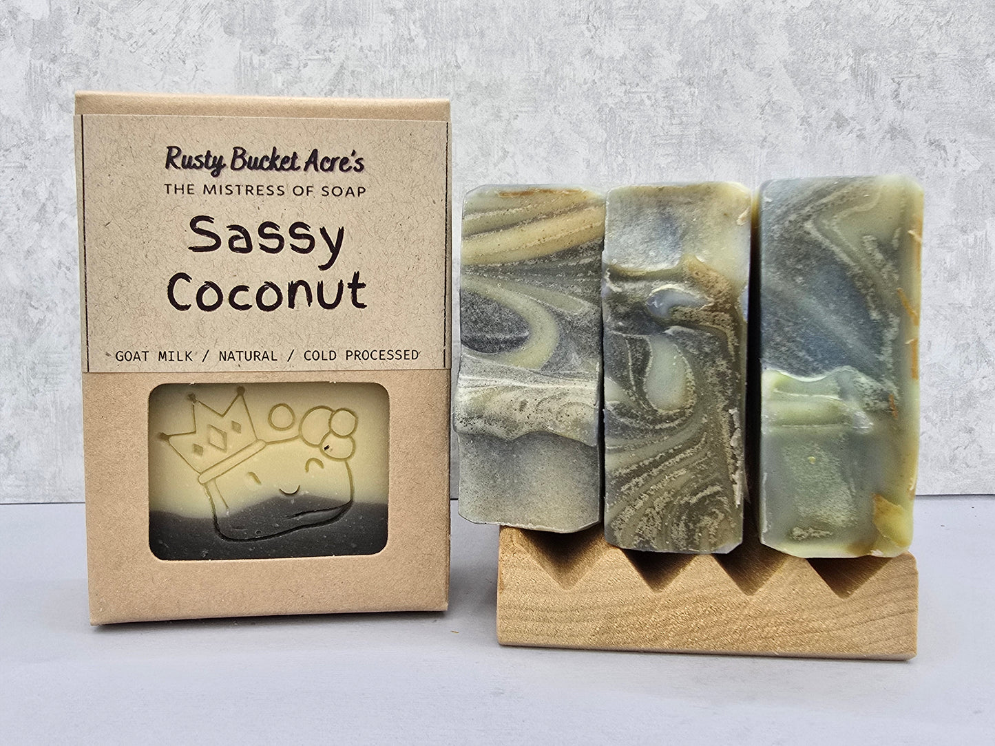 Three artisan soap bars with a marbled green, gray, and beige pattern displayed on a small wooden stand next to their kraft paper packaging box labeled 'Rusty Bucket Acre's Sassy Coconut'.