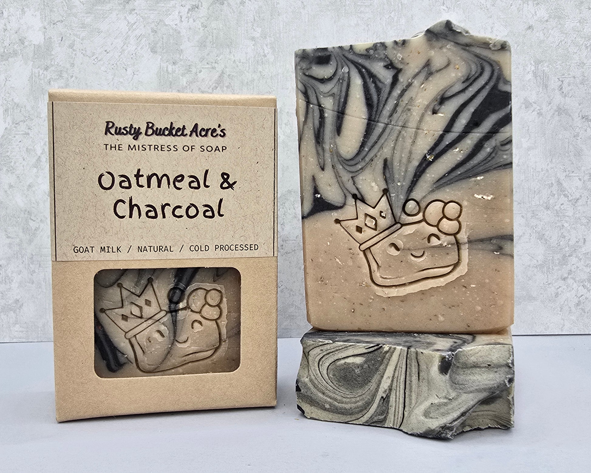 A handmade bar of soap with a carved crown design, shown next to its kraft paper packaging box. The soap has a marbled oatmeal and charcoal pattern, and the box features a clear window displaying the soap design.