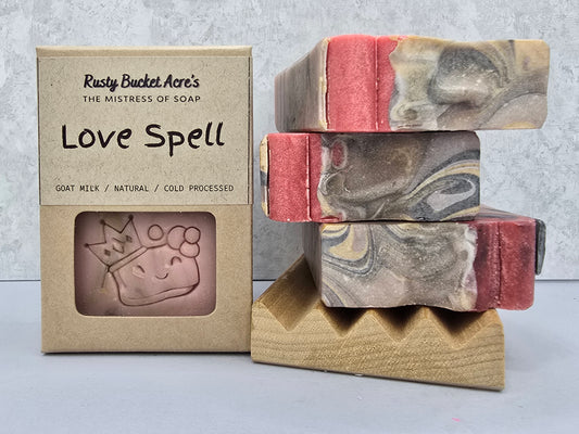 Three marbled handmade soap bars with pink and gray swirls are stacked on a wooden block next to their kraft paper packaging box labeled 'Love Spell' by Rusty Bucket Acre's.