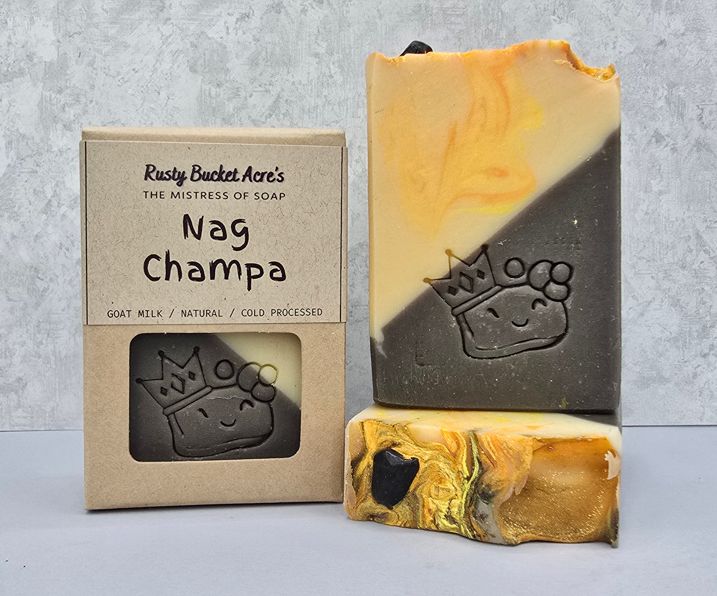 A handmade soap bar labeled 'Nag Champa' by Rusty Bucket Acre's, shown alongside its kraft paper packaging box. The soap has a diagonal split design of dark charcoal gray and warm marbled yellow-gold tones, with a carved crown logo.