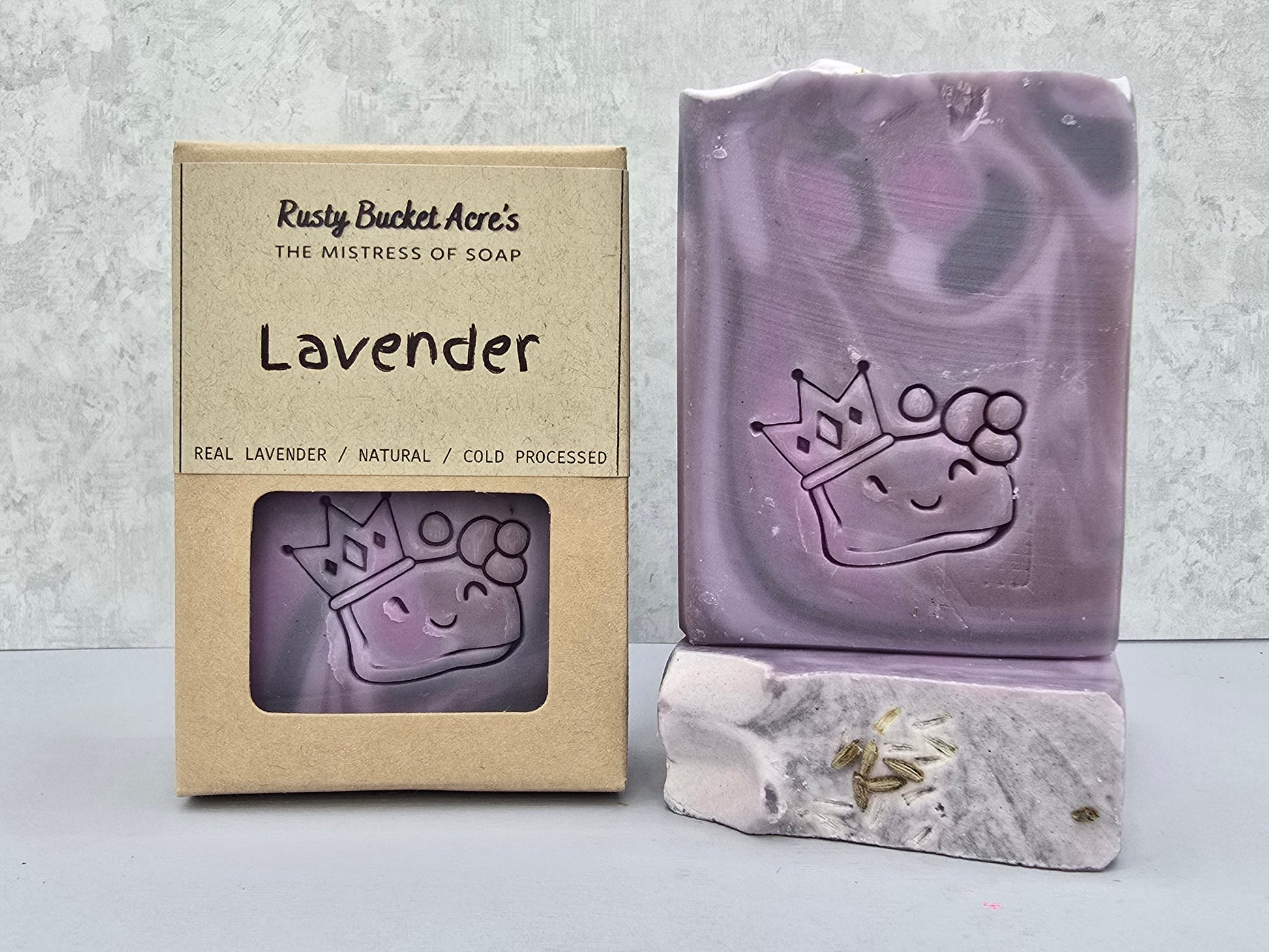 Lavender handcrafted soap bar with a crown design embossed on it, shown next to its kraft paper packaging box labeled 'Rusty Bucket Acre's Lavender'.