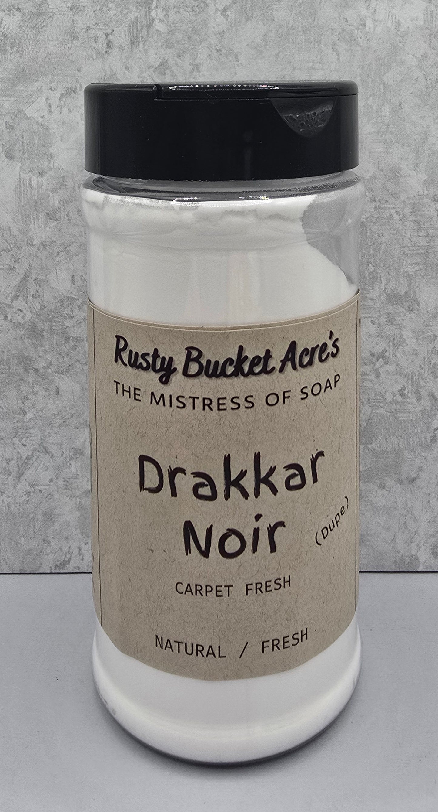 A clear plastic jar of Rusty Bucket Acre's Drakkar Noir carpet fresh soap powder, featuring a beige paper label and a black lid, set against a gray textured background.