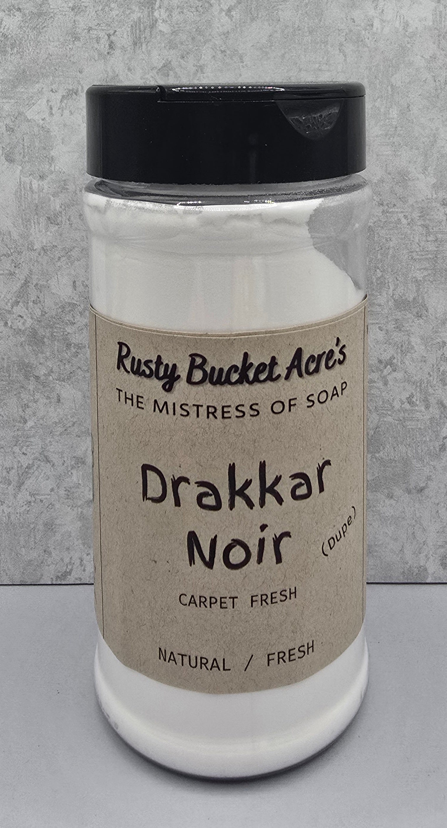 A clear plastic jar of Rusty Bucket Acre's Drakkar Noir carpet fresh soap powder, featuring a beige paper label and a black lid, set against a gray textured background.