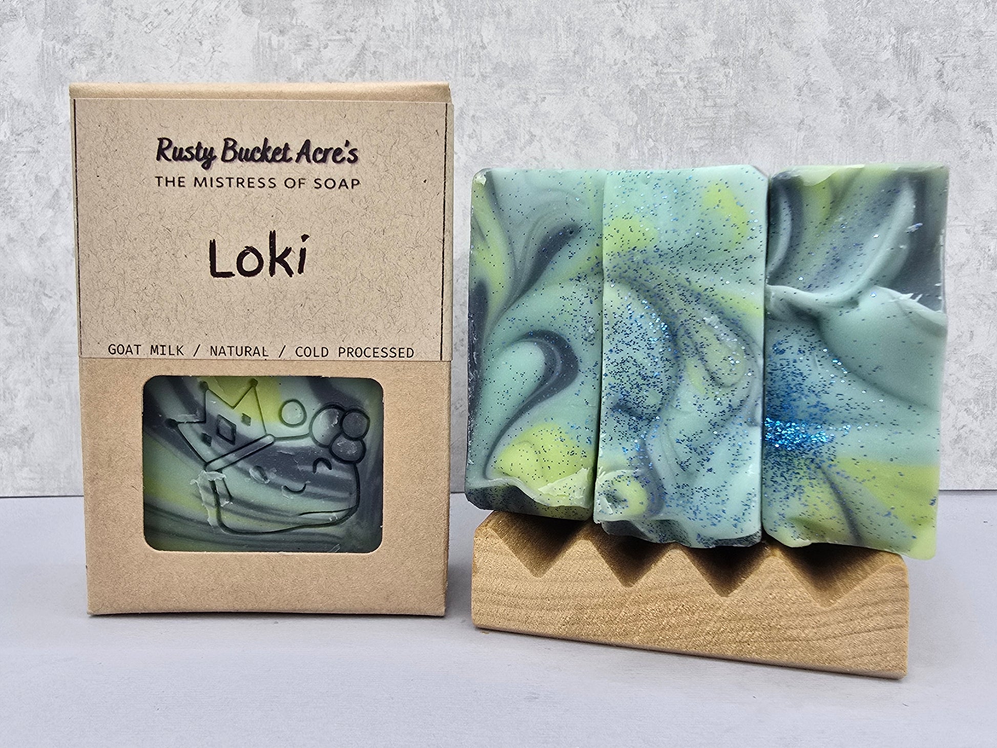 A handmade Loki soap bar with visible green, blue & yellow colors, displayed next to its kraft paper packaging box labeled 'Rusty Bucket Acre's Loki'.
