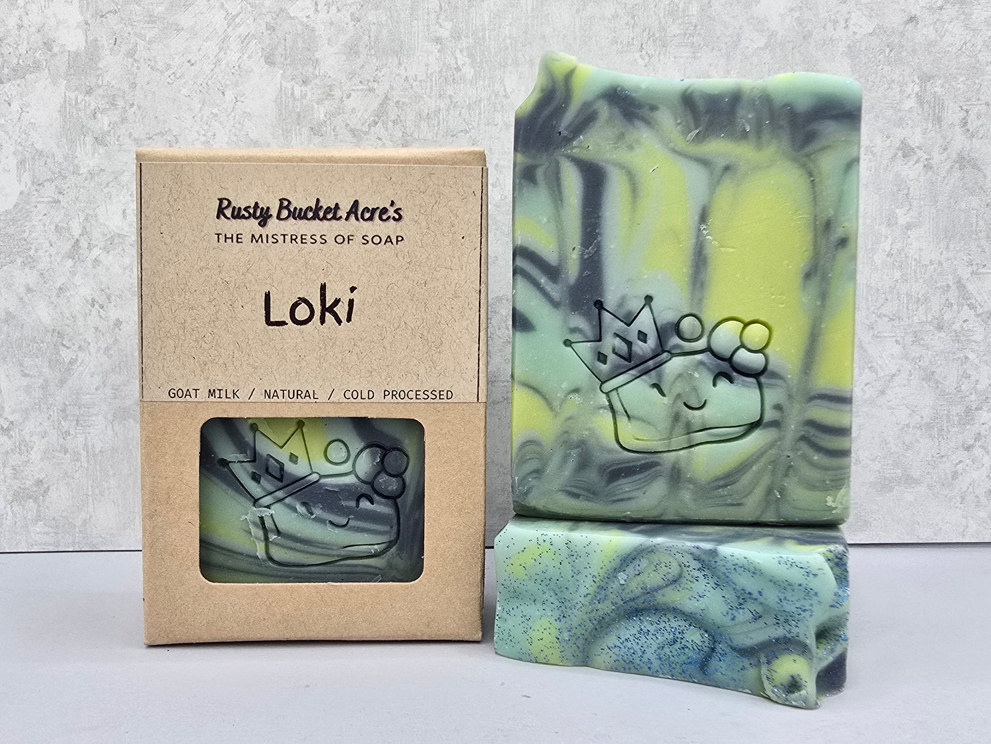 Loki handcrafted soap bar with a crown design embossed on it, shown next to its kraft paper packaging box labeled 'Rusty Bucket Acre's Loki'.