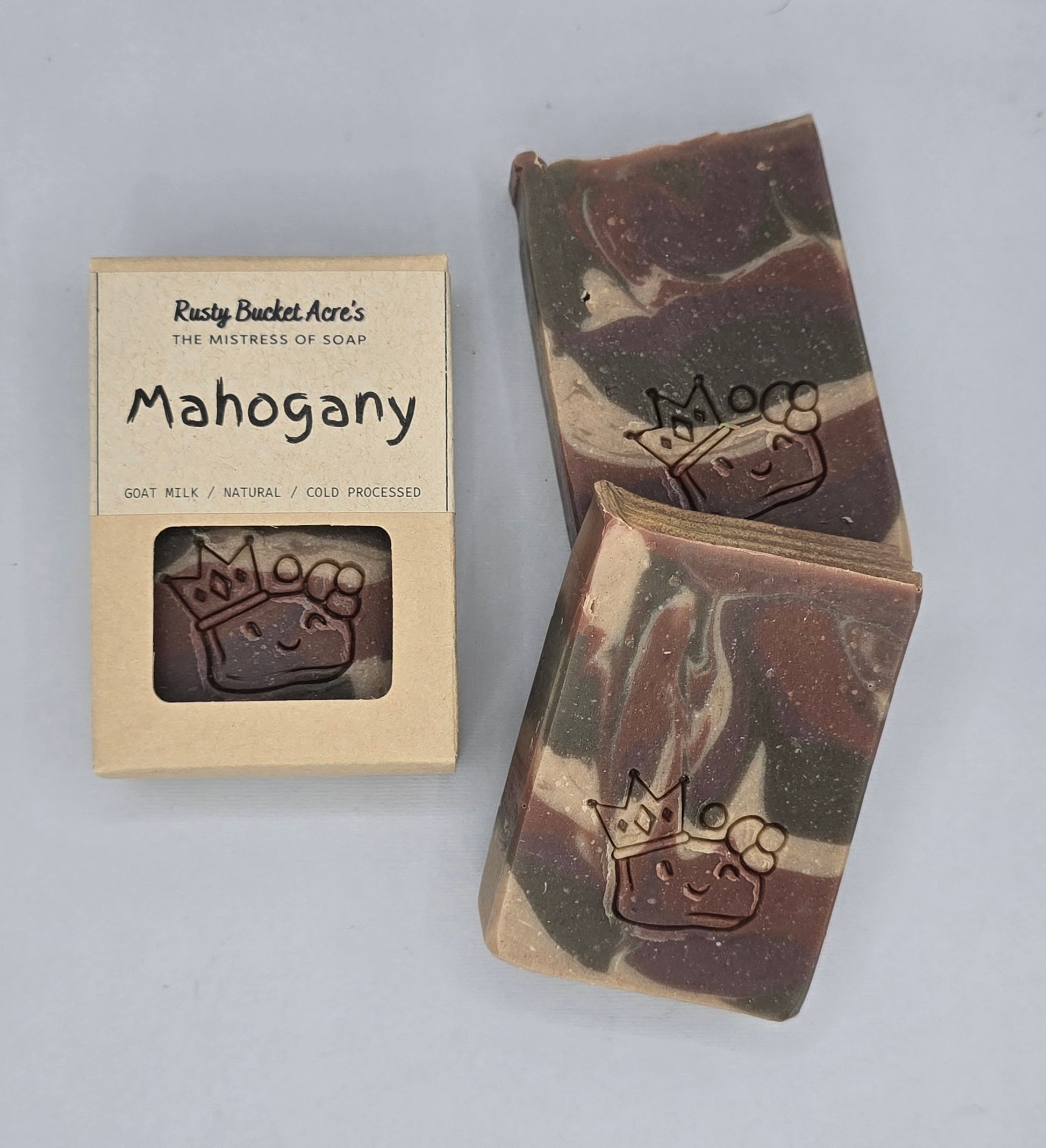 Two bars of handmade soap with a brown and cream marbled pattern, each embossed with a crown and flower design, displayed next to their kraft paper packaging box labeled 'Mahogany' by Rawly Bucket Acre's.