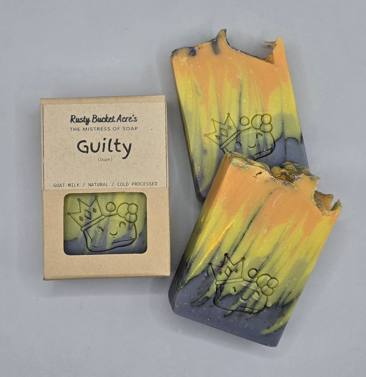 Two handmade goat milk soap bars with a yellow and dark gray marbled design, next to their kraft paper packaging box labeled 'Guilty' by Rusty Bucket Ate's.