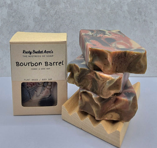 A handmade soap bar with a brown, beige, and black marbled camouflage pattern, shown next to its kraft paper packaging box labeled 'Rusty Bucket Acre's Bourbon Barrel'.