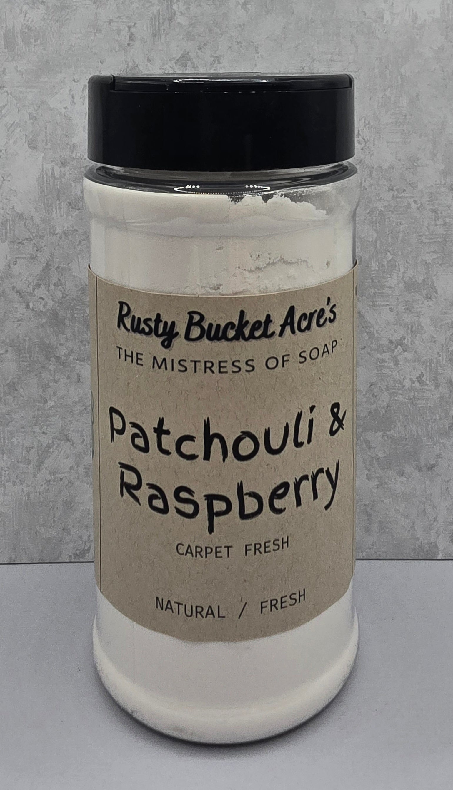 A clear plastic jar of Rusty Bucket Acre's Patchouli & Raspberry Carpet Fresh soap powder, featuring a kraft paper label and a black lid, set against a gray textured background.