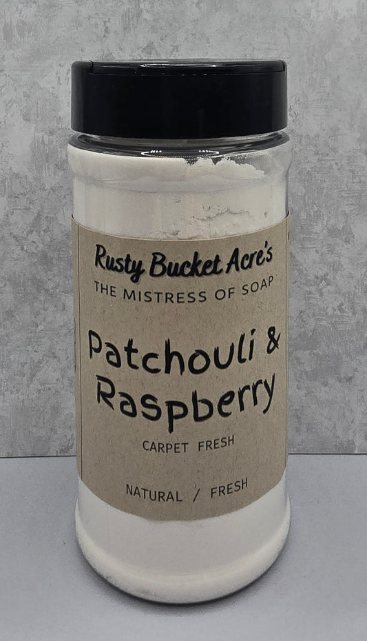 A clear plastic jar of Rusty Bucket Acre's Patchouli & Raspberry Carpet Fresh soap powder, featuring a kraft paper label and a black lid, set against a gray textured background.