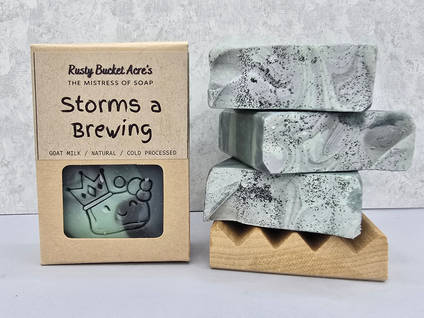 A boxed bar of soap next to three stacked soap bars on a wooden stand. The kraft paper box reads 'Rusty Bucket Acre's The Mistress of Soap Storms a Brewing' and shows a window with an embossed goat head.