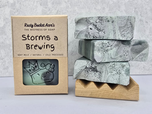 A boxed bar of soap next to three stacked soap bars on a wooden stand. The kraft paper box reads 'Rusty Bucket Acre's The Mistress of Soap Storms a Brewing' and shows a window with an embossed goat head.