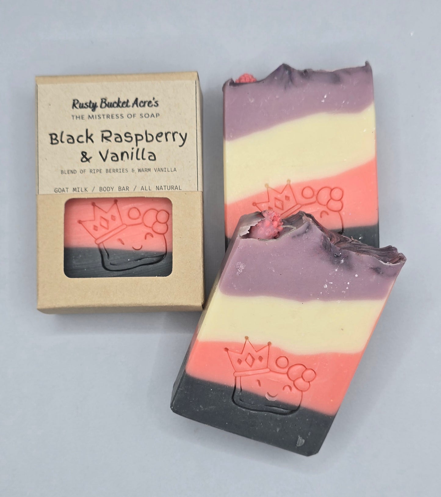 Two handmade soap bars and their kraft paper packaging box from Rusty Bucket Art's, featuring a black, cream, and pink striped design with a purple top layer and a crown embossed on the soaps.