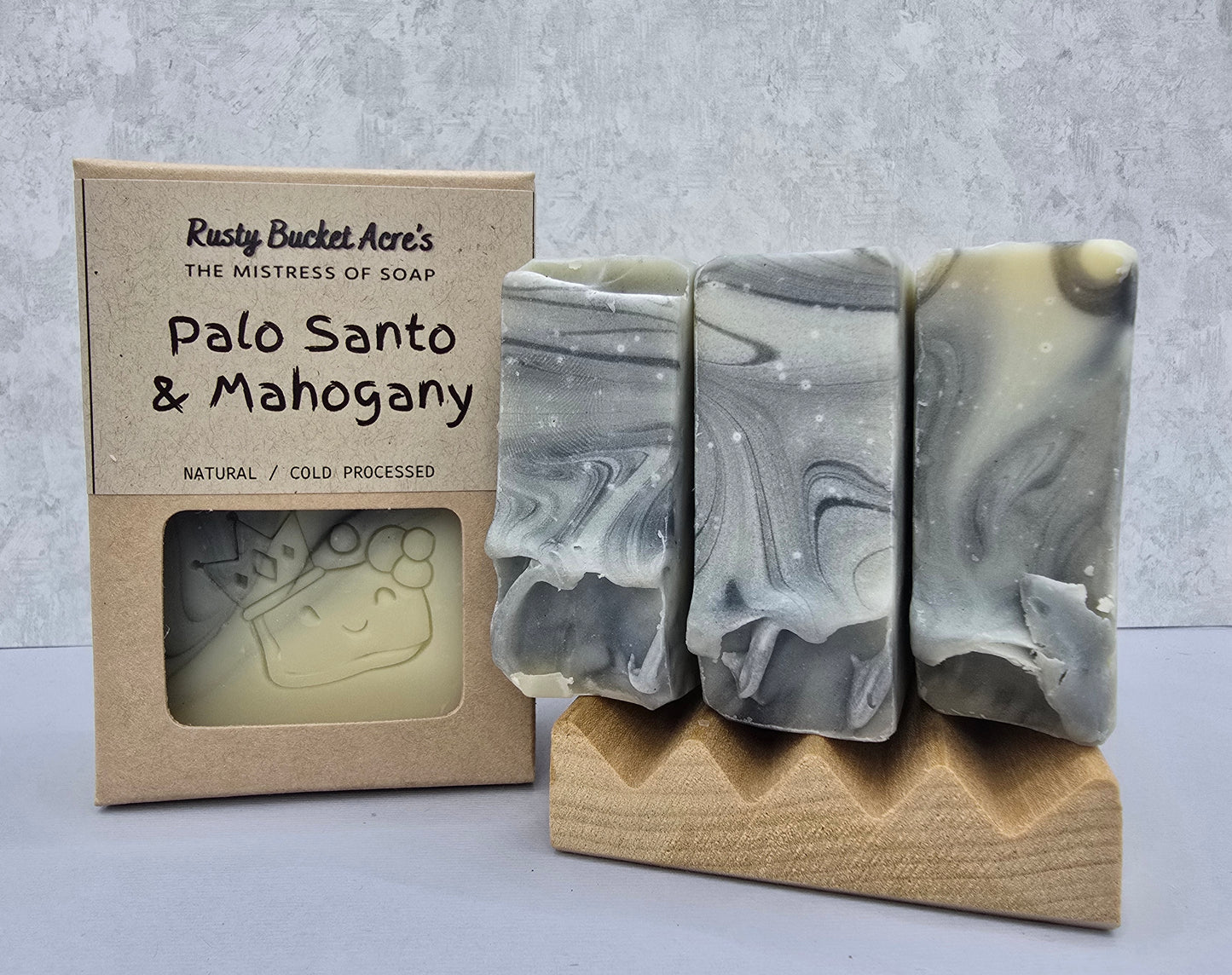Three handmade soap bars with gray, white, and beige marbled swirls displayed next to their kraft paper packaging box labeled 'Rusty Bucket Acre's Palo Santo & Mahogany'.