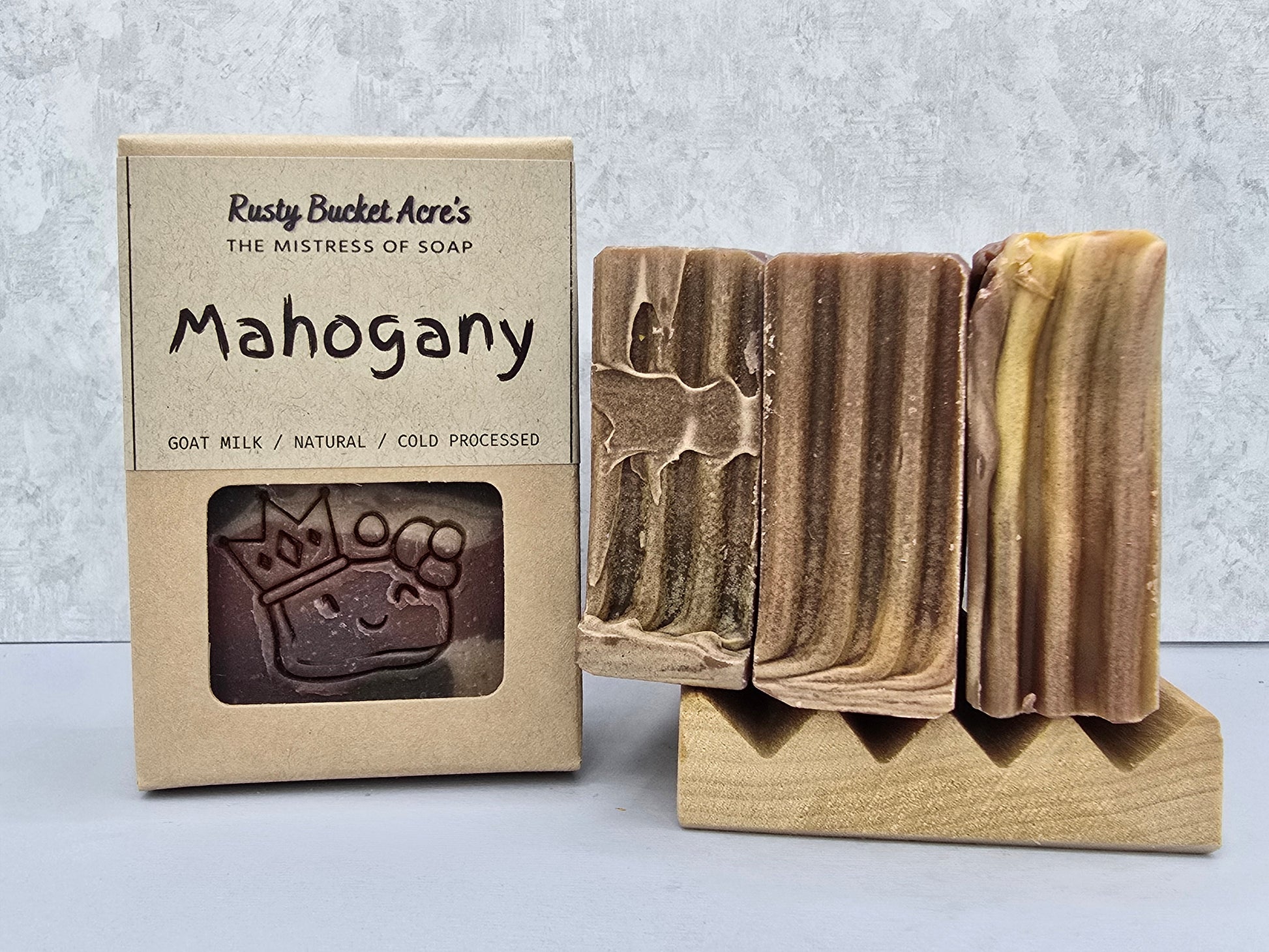 A bar of handmade soap labeled 'Mahogany' by Rusty Bucket Acre's, shown next to its kraft paper packaging box. The soap has a marbled brown and beige coloration and is displayed on a wooden stand against a light gray textured background.
