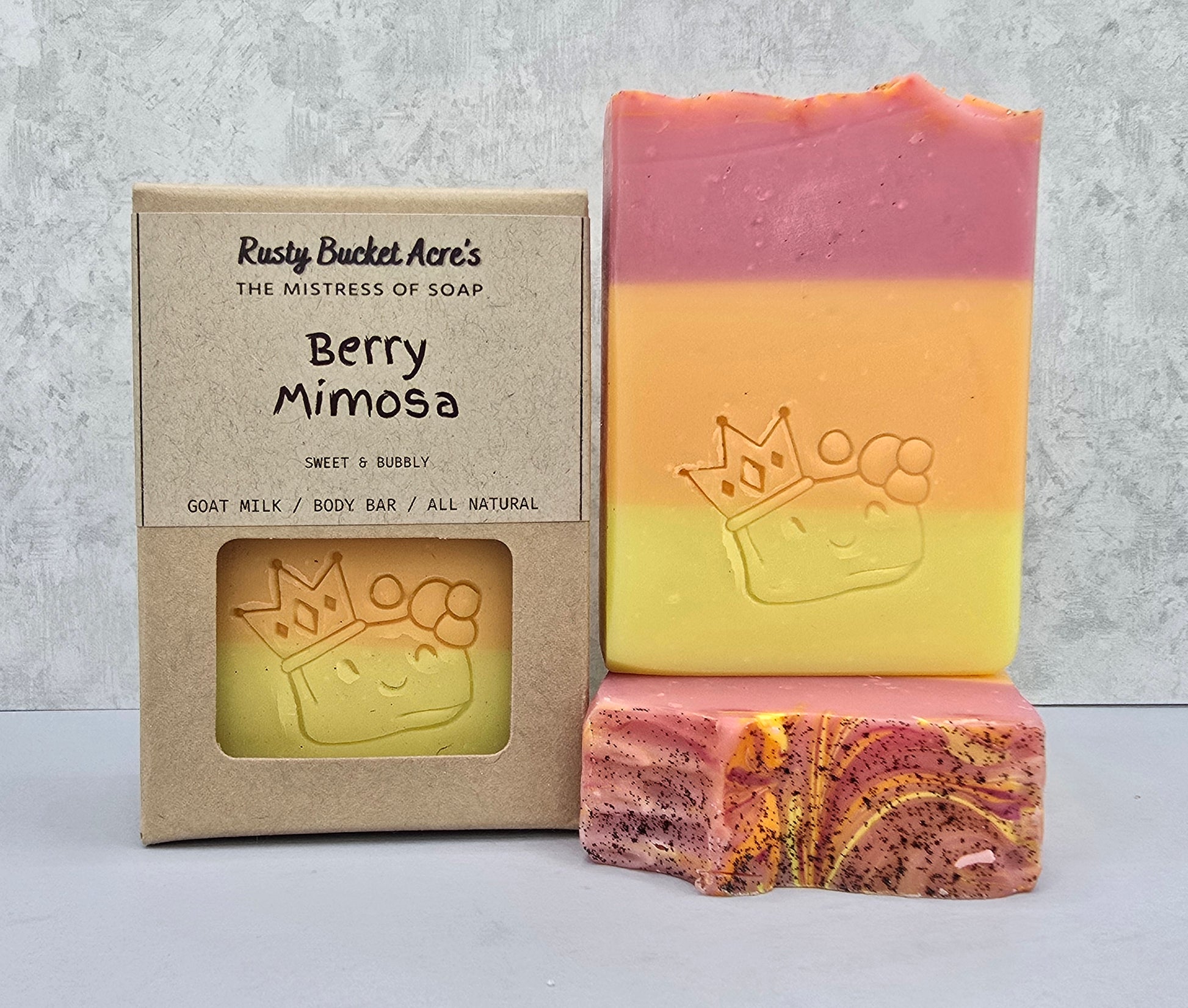 A handmade three-layer soap bar in pink, orange, and yellow with a carved crown and paw print, shown next to its kraft paper packaging box labeled 'Berry Mimosa' by Rusty Bucket Acre's, and a cut piece showing the marbled interior.