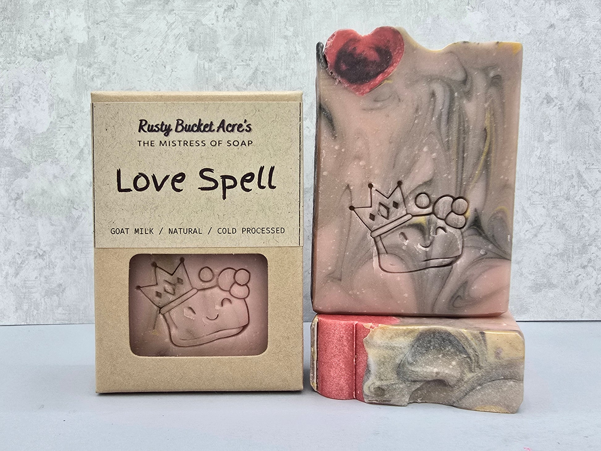 A handmade soap bar labeled 'Love Spell' by Rusty Bucket Acre's, shown next to its kraft paper packaging box and a smaller side view of the soap.